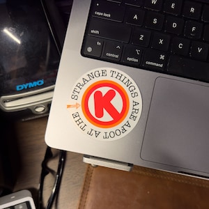 May include: A white sticker with a red circle and a white letter K in the center. The circle has an orange outline. The text around the circle reads "Strange Things Are Afoot At The"