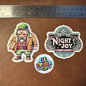 May include: Three stickers on a brown surface. One features a cartoon man with a large mustache holding a hot dog. Another says "Night of Joy, French Quarter." The third is a round sticker with "Lil Sticker Co."