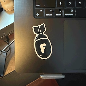 Puede incluir: Una pegatina en blanco y negro con forma de bomba con la letra "F" pegada a un portátil. La pegatina tiene un contorno blanco y está sobre una superficie negra del portátil. El teclado del portátil es visible.