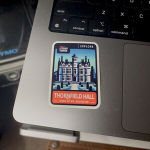 May include: A sticker with a blue and white illustration of Thornfield Hall, the fictional home of Mr. Rochester from the novel Jane Eyre. The sticker is on a laptop keyboard and has the text "Explore", "Literary Tourism", and "Home of Mr. Rochester".