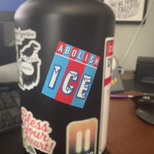 May include: Black water bottle covered in stickers, including one that says "ABOLISH ICE" in white text on a blue and red striped background. Other stickers include a raven, the words "Bless Your Heart!", and a pause symbol.