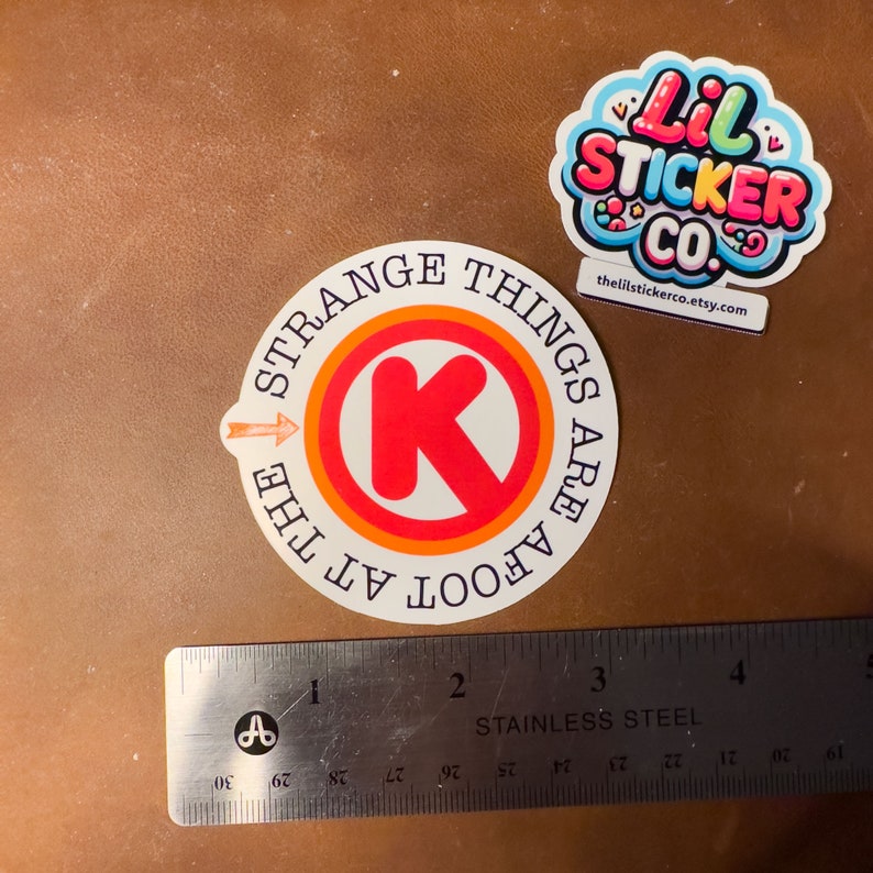 May include: A white sticker with a red "K" in a circle with orange and white rings. The text around the circle reads "STRANGE THINGS ARE AFOOT AT THE". The sticker is next to a ruler.