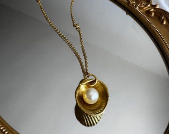 May include: A gold-colored necklace featuring a seashell pendant with a pearl. The pendant is detailed with textured ridges and hangs from a delicate chain. The necklace is displayed on a gold-framed mirror.