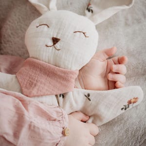 May include: A soft, white bunny plush toy with embroidered facial features and a pink neckerchief. The bunny's ears and paws are adorned with floral patterns. A child's hands gently hold the toy, suggesting a comforting and cuddly design.