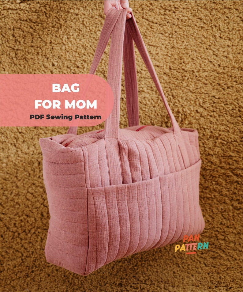 Może przedstawiać: R&oacute;żowa torba typu tote z pionowymi pikowanymi detalami i dwiema zewnętrznymi kieszeniami. Torba ma długie uchwyty, a widoczny jest tekst "BAG FOR MOM PDF Sewing Pattern". Torba jest trzymana na tle teksturowanego, brązowego tła.
