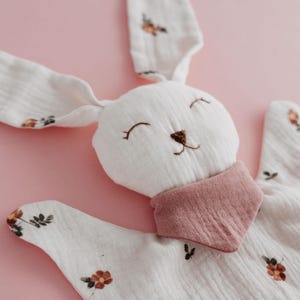 May include: A white bunny-shaped baby comforter with long ears and a pink bib. The comforter is decorated with small floral patterns in shades of orange and brown. The bunny's face is embroidered with a smiling expression.