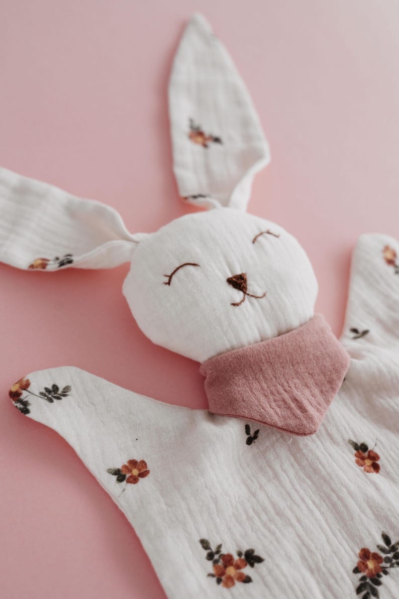 May include: A white bunny-shaped baby comforter with long ears and a pink bib. The comforter is decorated with small floral patterns in shades of orange and brown. The bunny's face is embroidered with a smiling expression.