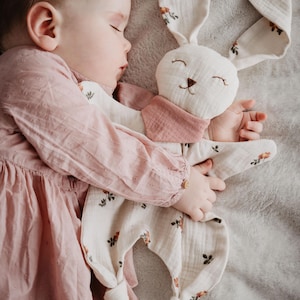 May include: A sleeping baby wearing a pink dress, cuddling a white bunny-shaped plush toy. The bunny has long ears, a pink neck wrap, and floral accents. The background is a soft, gray blanket.
