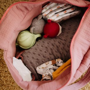 Może przedstawiać: R&oacute;żowa torba dla dziecka wypełniona niezbędnymi rzeczami dla niemowląt. W środku znajdują się pieluchy, szara szydełkowa zabawka, czerwona gumowa zabawka, zielony kubek niekapek, szary kocyk i ubranka dla dziecka. Torba jest otwarta, ukazując zawartość.