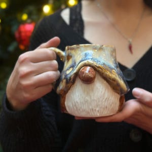 Puede incluir: Una taza de cerámica con forma de gnomo con nariz marrón, barba blanca y parte superior en forma de tejado. La taza tiene un asa dorada y se sostiene frente a un árbol de Navidad con adornos rojos y dorados. El diseño de la taza es caprichoso y festivo.