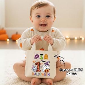 May include: A beige tote bag with the text "My 1st Halloween" and a mummy, ghost, and pumpkin graphic. The bag is held by a baby wearing a cream-colored sweater with pumpkin designs.