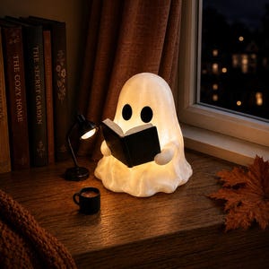 May include: A white, glowing ghost-shaped lamp holding a small black book, positioned next to a miniature black desk lamp, a tiny black mug, and a single dried maple leaf. The ghost has two black oval eyes and emits a warm, soft light.
