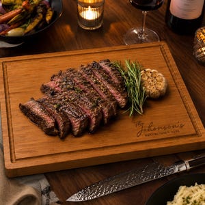 May include: A rectangular wooden chopping board with a juice groove, featuring the engraved text The Johnson's ESTABLISHED 2026. The board holds a sliced seared steak, a sprig of fresh rosemary, and a roasted garlic bulb on its surface.