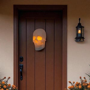 May include: A glowing white skull decoration hangs on a brown wooden door. The skull has orange glowing eyes and is surrounded by a fall-themed porch display with pumpkins, orange flowers, and a black lantern.