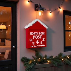 以下が含まれることがあります： 白い屋根と雪の結晶が特徴の赤い「North Pole Post」の郵便ポスト。 郵便ポストは灰色の壁に取り付けられており、「North Pole Post」と「The Campbell Family」の文字が白で表示されています。 ライトのストリングと黒い照明器具が郵便ポストの上にあります。