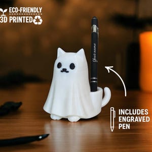 Könnte beinhalten: Ein weißer, katzenförmiger Stifthalter mit geisterhaftem Design, der einen schwarzen Stift hält. Auf dem Stift steht "Your name". Der Halter ist 3D-gedruckt und umweltfreundlich. Der Text "Includes engraved pen" ist ebenfalls enthalten.