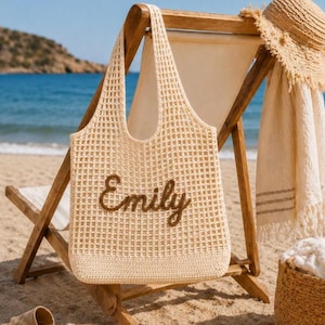 Personalized crochet beach tote bag with the name Emily embroidered in brown lettering, styled on a beach chair by the ocean with a straw hat and summer accessories. Neutral cotton mesh shoulder bag perfect for beach vacations, bridesmaid gifts, coastal aesthetic outfits, pool days, and personalized summer gifts for women.