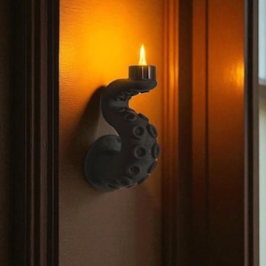May include: A black octopus tentacle-shaped candle holder fixed to a wooden wall. The tentacle curves upwards, supporting a lit candle. The warm lighting creates a cosy atmosphere.