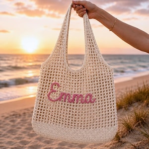 Personalized crochet beach tote bag with the name Emma in pink lettering, photographed at sunset by the ocean. Neutral cotton mesh shoulder bag with a coastal aesthetic, perfect for beach vacations, bridesmaid gifts, summer travel, pool days, and personalized gift ideas for women.