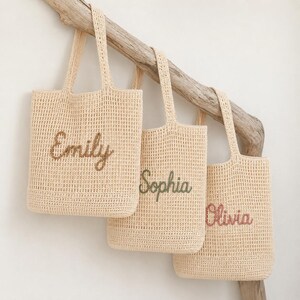 May include: Three beige crochet tote bags with embroidered names: "Emily" in brown, "Sophia" in green, and "Olivia" in pink. The bags hang from a wooden branch against a white background. Each bag has a rectangular shape and sturdy handles.