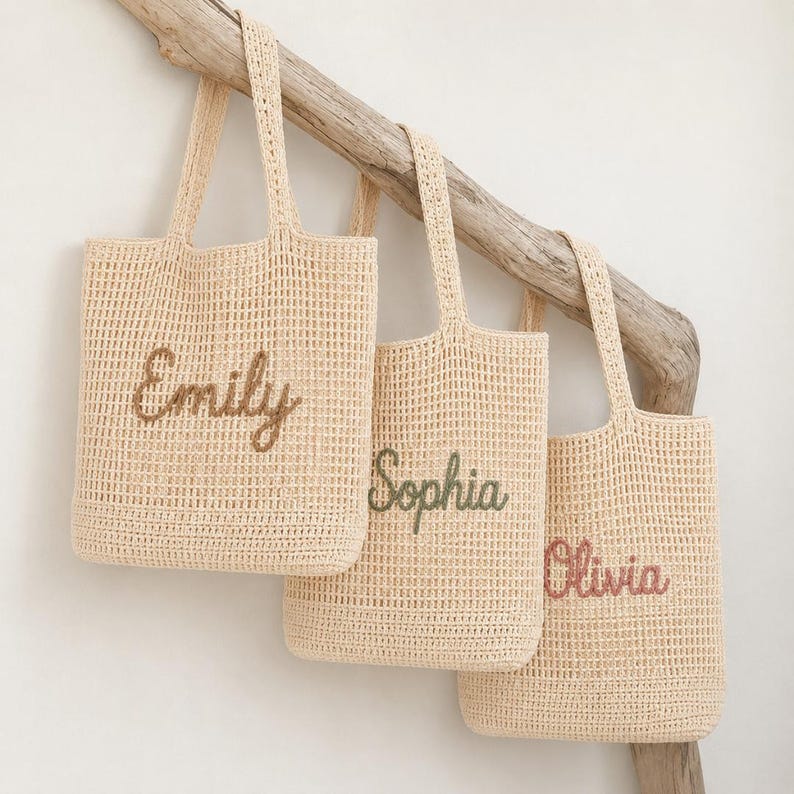 May include: Three beige crochet tote bags with embroidered names: "Emily" in brown, "Sophia" in green, and "Olivia" in pink. The bags hang from a wooden branch against a white background. Each bag has a rectangular shape and sturdy handles.