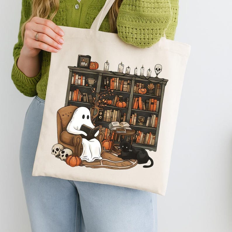 Op de afbeelding: Een witte tote bag met een griezelige Halloween-illustratie. Een geest zit op een stoel en leest een boek, een zwarte kat zit op de vloer en een boekenkast is gevuld met boeken en pompoenen.