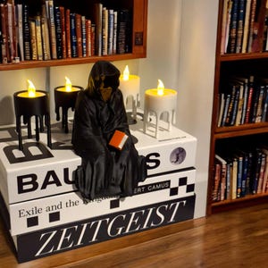May include: A black hooded figure sits on a stack of books with the titles "Bauhaus", "Exile and the Kingdom", and "Zeitgeist".  The figure is holding a book and there are three candle holders with lit candles on the table.