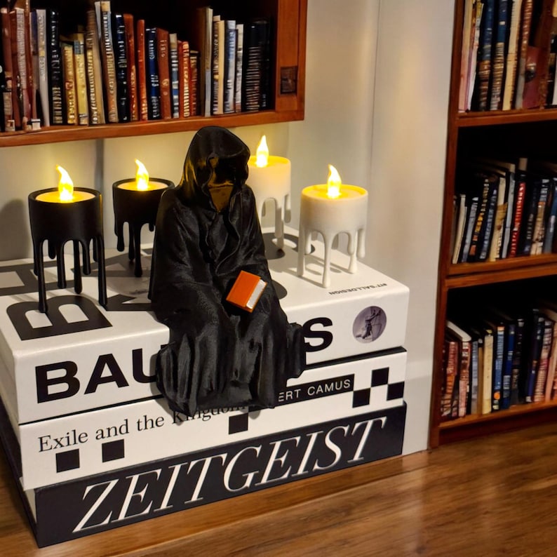 May include: A black hooded figure sits on a stack of books with the titles "Bauhaus", "Exile and the Kingdom", and "Zeitgeist".  The figure is holding a book and there are three candle holders with lit candles on the table.