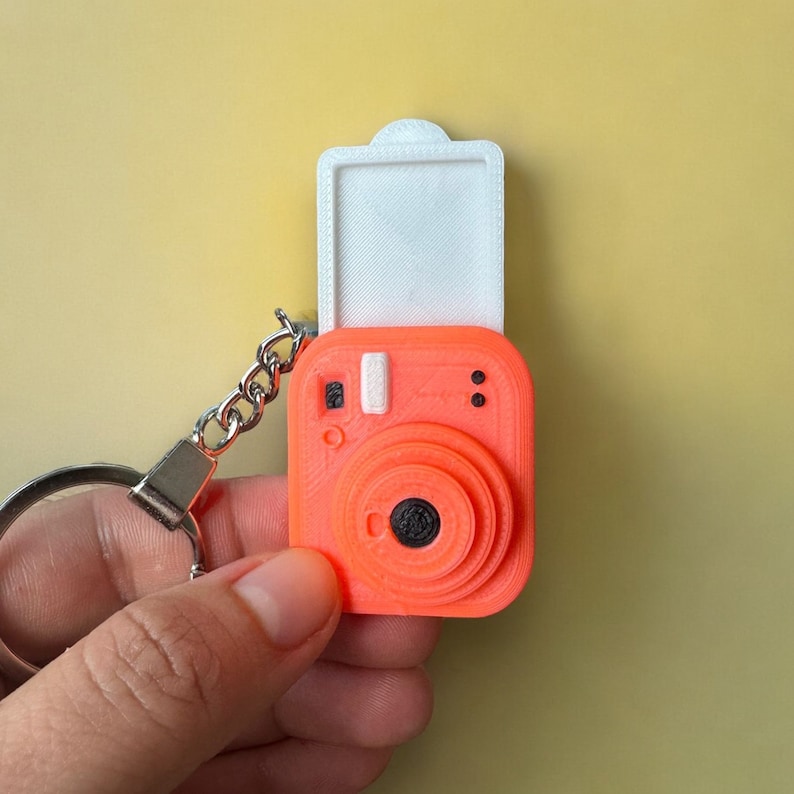 May include: A bright orange camera keyring with a white lens cover. The camera has a black lens and a silver key ring.