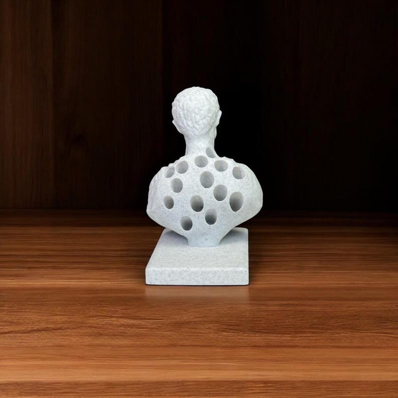 May include: White ceramic bust sculpture with a hollowed-out back featuring multiple circular holes. The bust is on a white base.