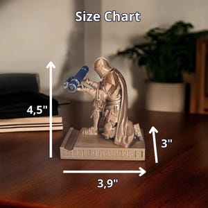 Puede incluir: Figura de Mandaloriano de color bronce, arrodillada y sosteniendo un telescopio azul. La base mide 9,9 cm de largo y 7,6 cm de ancho. La figura mide 11,4 cm de alto. El texto "Size Chart" est&aacute; arriba.