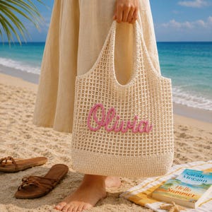 May include: A cream-coloured, open-weave tote bag with the name "Olivia" embroidered in pink. The bag is held on a sandy beach with sandals and a book visible. The ocean and sky are in the background.