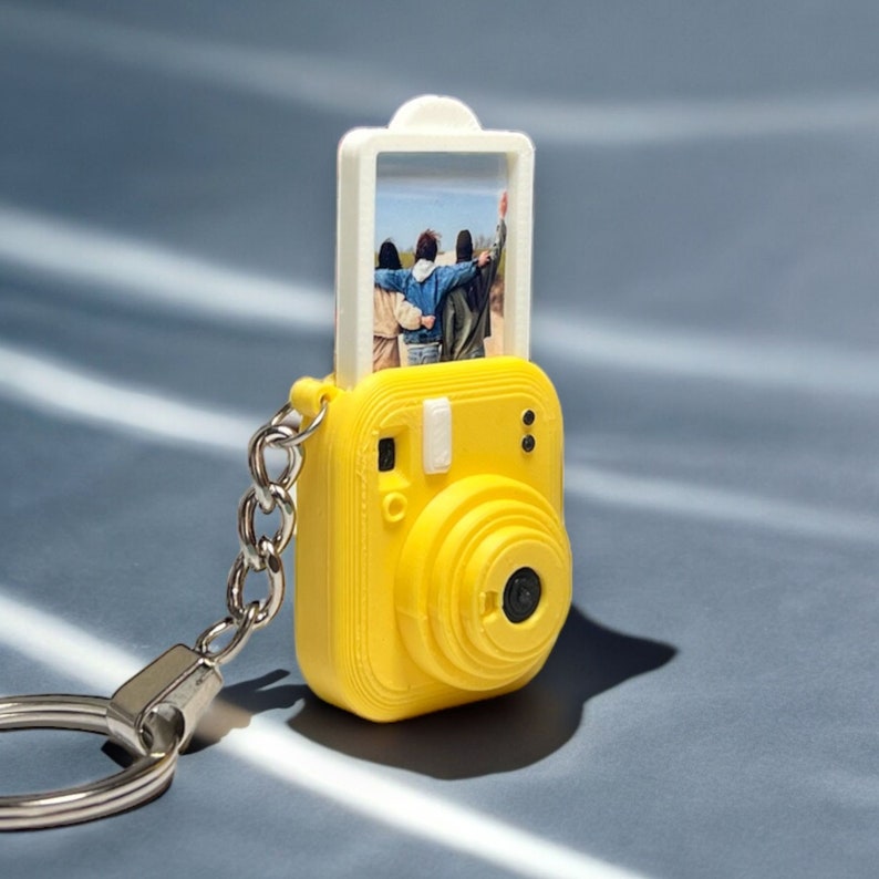 May include: A yellow plastic keychain shaped like an instant camera with a clear plastic insert for a photo. The photo shows three people standing together. The keychain has a silver metal ring.
