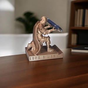 Puede incluir: Soporte para bol&iacute;grafo con forma de caballero de color bronce arrodillado y sosteniendo un bol&iacute;grafo azul. El caballero lleva una armadura completa, con capa y casco. El soporte para bol&iacute;grafo est&aacute; sobre una base rectangular.