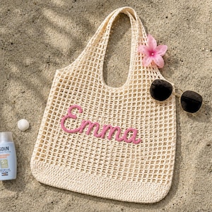 May include: A cream-coloured crocheted tote bag with the name "Emma" in pink script. The bag is on a sandy surface, with sunglasses and a pink flower. A bottle of sunscreen and a small white shell are also visible.
