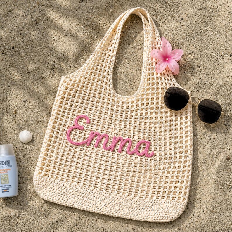 May include: A cream-coloured crocheted tote bag with the name "Emma" in pink script. The bag is on a sandy surface, with sunglasses and a pink flower. A bottle of sunscreen and a small white shell are also visible.