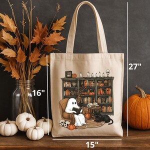 Op de afbeelding: Een beige tote bag met een geest die een boek leest voor een boekenkast. De geest is wit en houdt een boek vast. Er zit een zwarte kat op de vloer voor de boekenkast. De tas is 15 inch breed en 27 inch hoog.