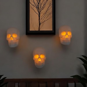 Puede incluir: Tres decoraciones de pared con forma de calavera blanca con ojos y narices de color naranja brillante están montadas en una pared gris claro. Una imagen enmarcada de un árbol está encima de las calaveras. Plantas verdes en primer plano.