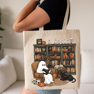 Op de afbeelding: Een witte tote bag met een geest die in een stoel leest voor een boekenkast vol boeken. De boekenkast is versierd met pompoenen, kaarsen en een schedel. De geest draagt een wit laken en heeft een zwart boek in zijn handen. De stoel is bruin en er zit een zwarte kat op de armleuning. De tote bag is een geweldige manier om je boeken en andere benodigdheden te dragen.