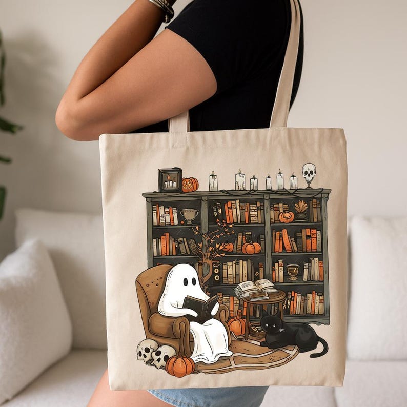 Op de afbeelding: Een witte tote bag met een geest die in een stoel leest voor een boekenkast vol boeken. De boekenkast is versierd met pompoenen, kaarsen en een schedel. De geest draagt een wit laken en heeft een zwart boek in zijn handen. De stoel is bruin en er zit een zwarte kat op de armleuning. De tote bag is een geweldige manier om je boeken en andere benodigdheden te dragen.