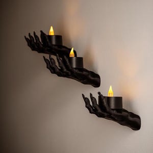May include: Three black hand-shaped wall sconces, each holding a black candle with a flickering yellow LED light. The hands are detailed with long, sharp fingernails, creating a spooky Halloween decoration.