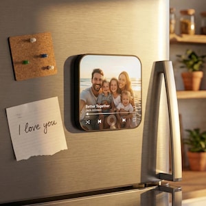 May include: A square, black magnetic photo frame displaying a family photo on a refrigerator. The photo shows a family of five at the beach. A corkboard with push pins and a note that says "I love you" are also on the fridge.
