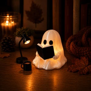 May include: A translucent white ghost figurine illuminated from within, holding an open black book. Beside the ghost is a small black desk lamp with a warm glowing light and a tiny black mug. The ghost has two solid black oval eyes.
