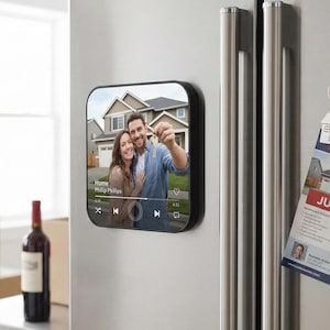 May include: A black square magnet with rounded corners, attached to a stainless steel refrigerator. The magnet displays a photo of a couple holding keys in front of a house, with the text "Home Phillip Phillips".