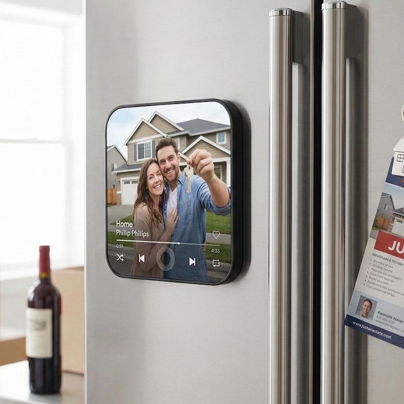 May include: A black square magnet with rounded corners, attached to a stainless steel refrigerator. The magnet displays a photo of a couple holding keys in front of a house, with the text "Home Phillip Phillips".