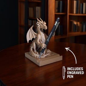 Puede incluir: Un portal&aacute;pices de drag&oacute;n de color bronce con un bol&iacute;grafo grabado. El drag&oacute;n est&aacute; posado sobre una base rectangular, sosteniendo el bol&iacute;grafo. El bol&iacute;grafo es negro con detalles plateados y tiene la inscripci&oacute;n "Your name".