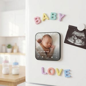 May include: A white refrigerator door displays colorful magnetic letters spelling "BABY" and "LOVE". A photo of a sleeping infant is on a black square magnet. An ultrasound image is also attached with a round magnet. Baby-themed decor.
