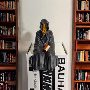 May include: A black hooded figure with a yellow face sits on a stack of books. The figure is holding a small brown book. The books are titled "Bauhaus", "The Kingdon", and "Other"