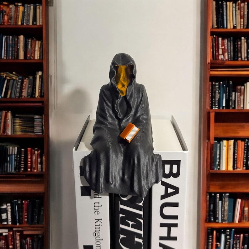 May include: A black hooded figure with a yellow face sits on a stack of books. The figure is holding a small brown book. The books are titled "Bauhaus", "The Kingdon", and "Other"