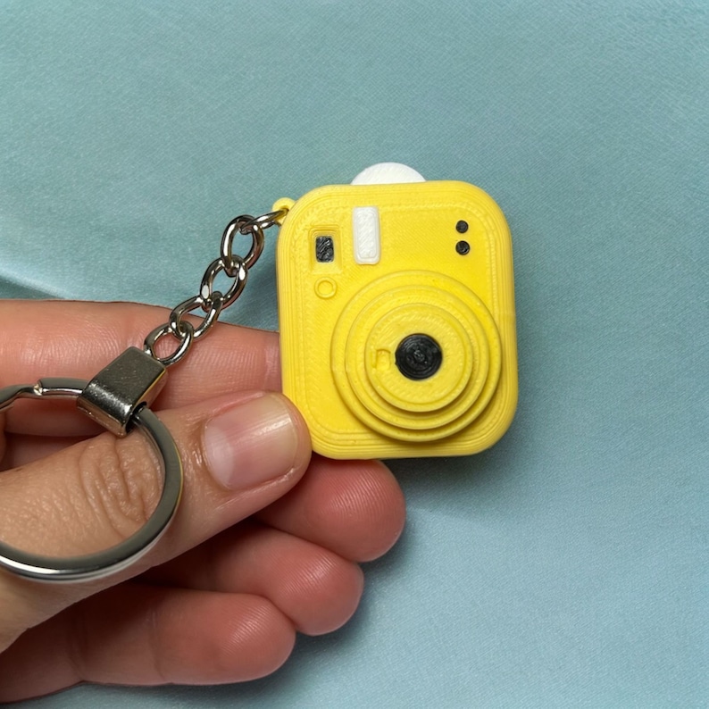 May include: A yellow plastic camera keyring with a silver key ring. The camera has a black lens and a white flash.